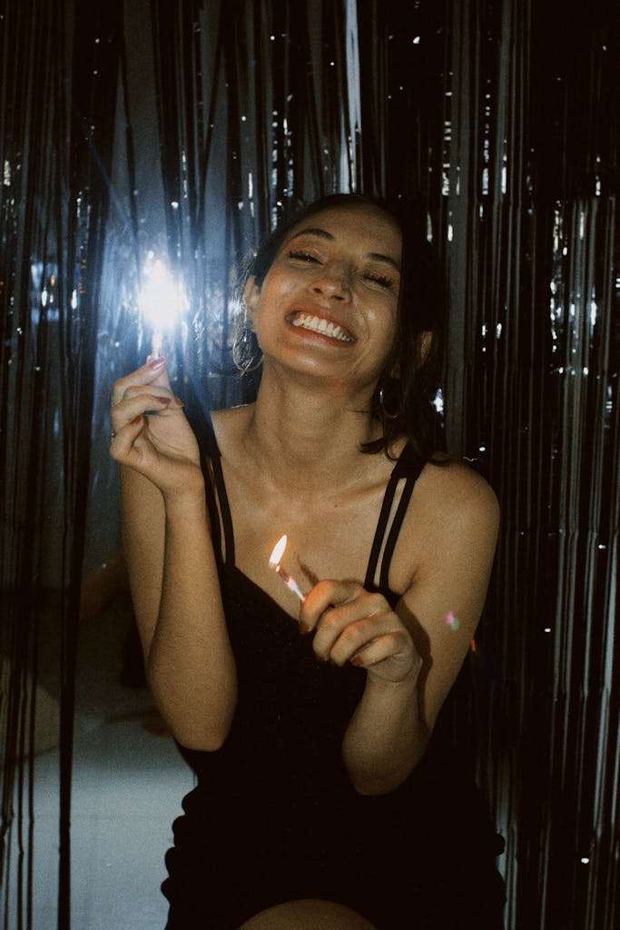 A cheerful woman in a black dress celebrates with candles and sparklers, exuding happiness.